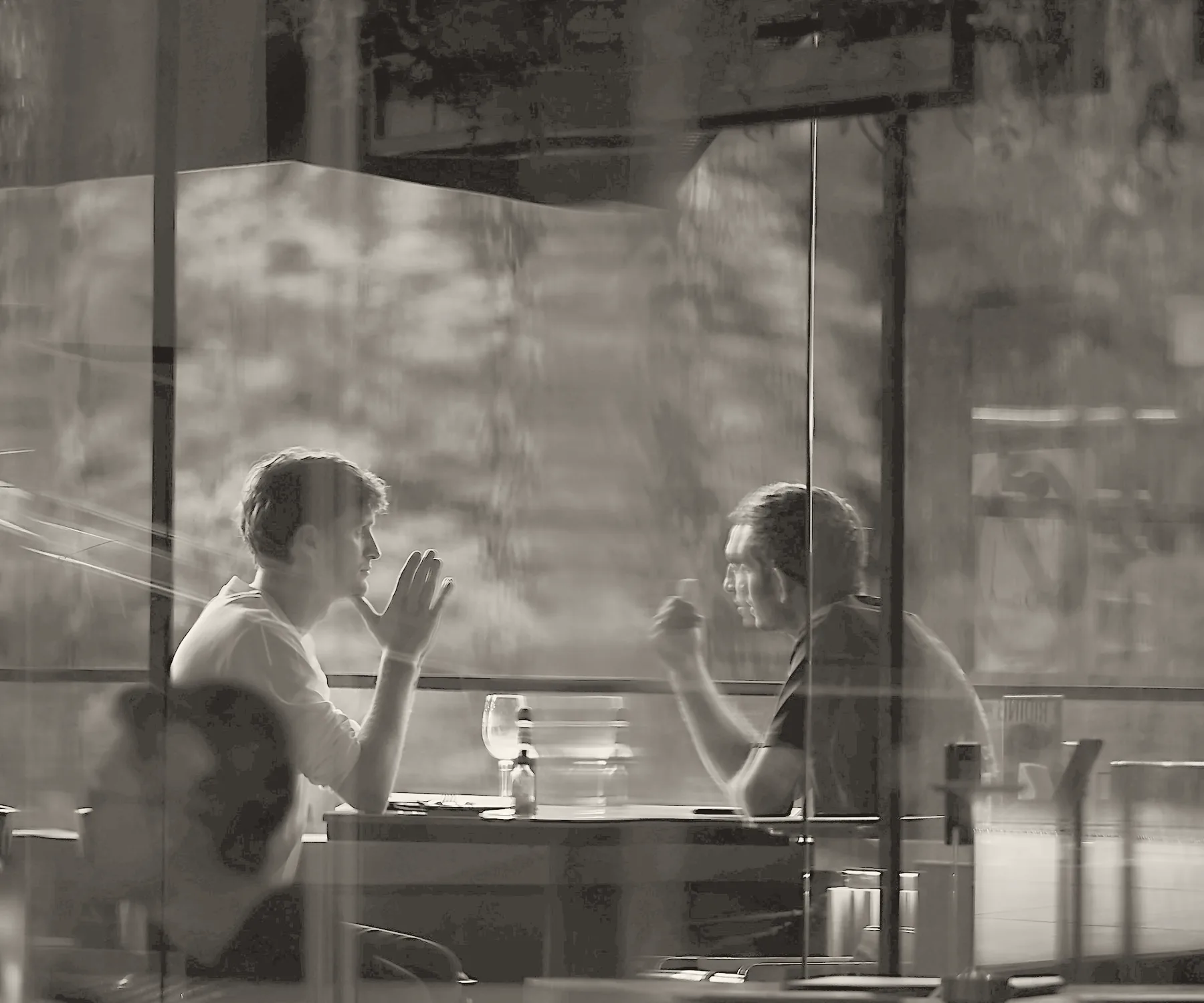 Two people talking across a café table, seen through the window